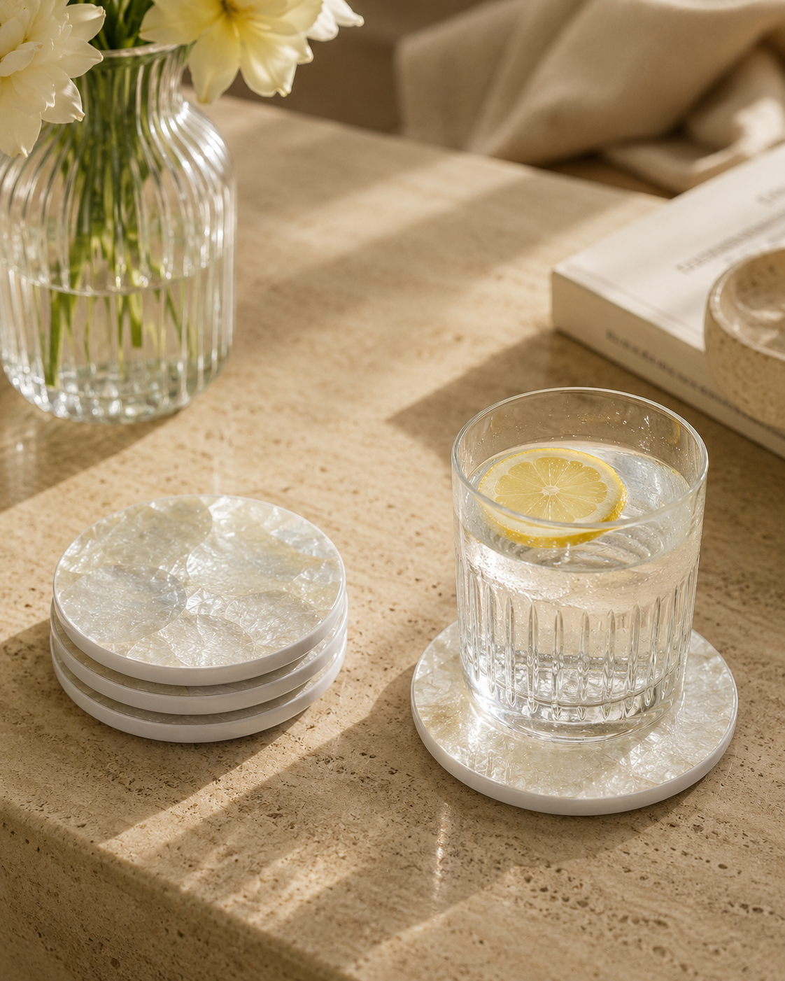 Glass of water with a lemon slice on a golden mother of pearl coaster, next to a vase with flowers on a wooden surface.